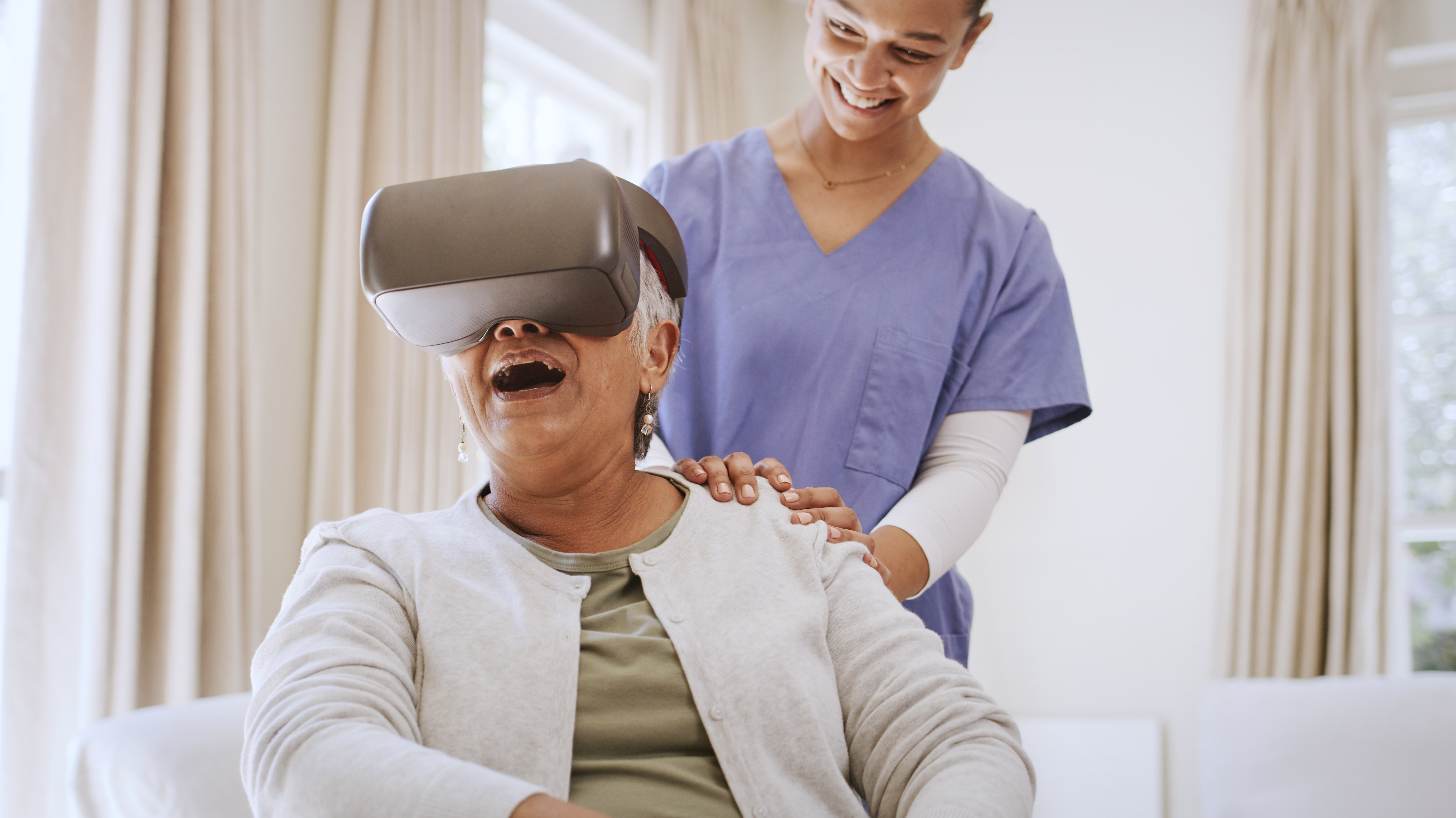 Older woman seated in care wearing virtual reality goggles with a nurse beside her